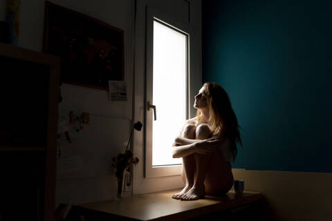 Serious young woman sitting on windowsill at home looking out stock photo