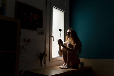 Seriöse junge Frau sitzt auf der Fensterbank zu Hause und hält eine Blume - AFVF05946