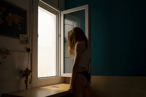 Junge Frau steht am Fenster zu Hause, lizenzfreies Stockfoto