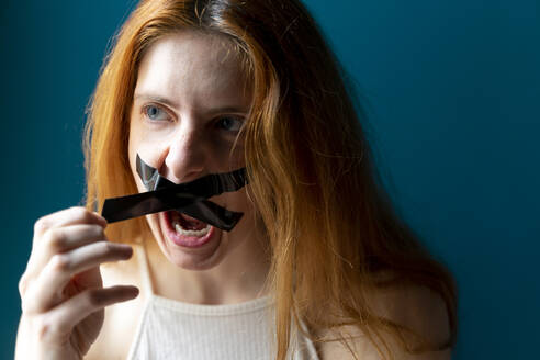 Portrait of screaming young woman pulling off sticky tape from her mouth - AFVF05955