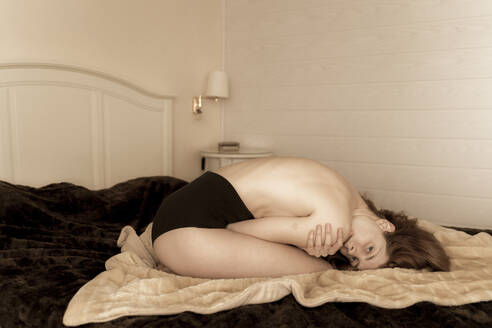 Junge Frau mit nacktem Oberkörper, die zu Hause auf dem Bett kauert - AFVF05961