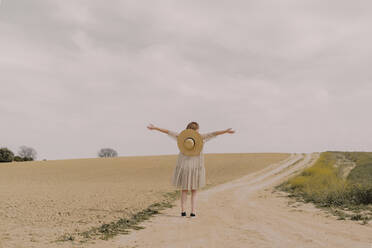 Woman with straw hat and vintage dress alone at a remote field road in the countryside - ERRF03069
