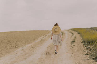 Frau mit Strohhut und Vintage-Kleid geht auf einem abgelegenen Feldweg auf dem Lande spazieren - ERRF03080