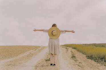 Rückansicht einer Frau mit Strohhut und Vintage-Kleid auf einem abgelegenen Feldweg auf dem Lande - ERRF03081