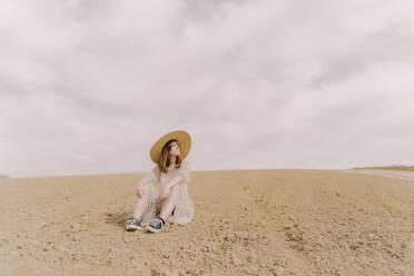 Frau mit Strohhut und Vintage-Kleid sitzt auf einem abgelegenen Feld auf dem Lande - ERRF03086