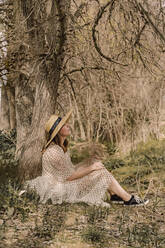 Frau mit Strohhut und Vintage-Kleid sitzt an einem Baum auf dem Lande - ERRF03102