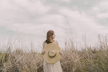 Woman in vintage dress holding straw hat at a remote field in the countryside - ERRF03106