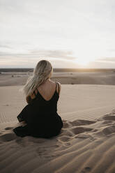 Rückenansicht einer blonden Frau, die auf einer Sanddüne sitzt und den Sonnenuntergang beobachtet, Algodones Dunes, Brawley, USA - LHPF01223