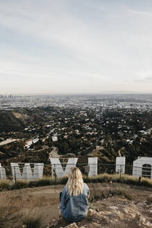 Rückenansicht einer blonden Frau, die hinter dem Hollywood-Schild sitzt und auf die Aussicht schaut, Beverly Hills, USA - LHPF01235
