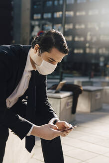 Businessman with face mask sitting outside using phone - MASF17327