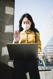 Businesswoman with face mask waving during video chat - MASF17328