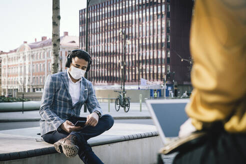 Businessman wearing face mask sitting outside using smartphone - MASF17329