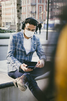 Businessman with smartphone sitting outside with face mask - MASF17330