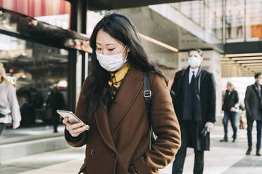 Frau mit Gesichtsmaske geht auf der Straße und benutzt ein Smartphone - MASF17345