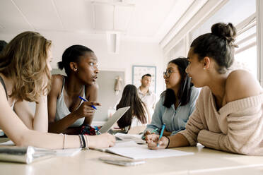 Female teenagers discussing while sitting by table in classroom - MASF17377