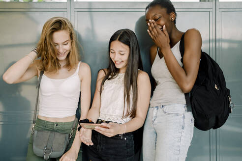 Lächelnde Mittelschülerin mit einem Freund, der ein Mobiltelefon benutzt, während sie im Schulkorridor stehen - MASF17389