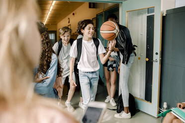 Verspielte männliche Schüler, die während der Mittagspause im Schulkorridor mit einem Basketball herumlaufen - MASF17398