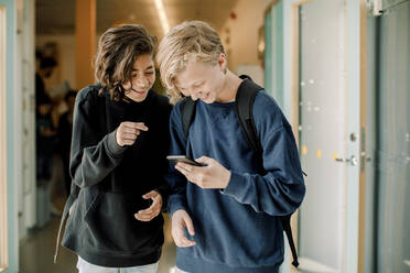 Lächelnde männliche Schüler, die im Schulkorridor ein Smartphone benutzen - MASF17402