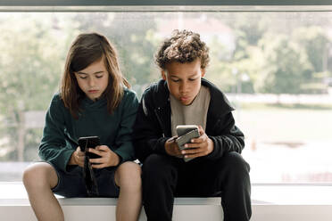 Männliche und weibliche Schüler, die ein Mobiltelefon benutzen, während sie an einem Glasfenster in der Schule sitzen - MASF17404