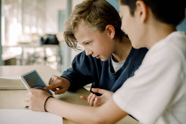 Männlicher Schüler, der auf ein digitales Tablet zeigt, während er mit einem Freund im Klassenzimmer sitzt - MASF17414