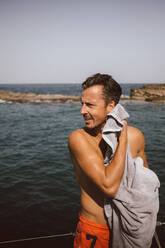 Mann schaut beim Abtrocknen mit Handtuch am Strand gegen den Himmel an einem sonnigen Tag weg - MASF17424