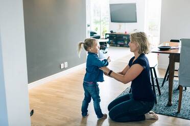 Side view of mother dressing daughter while kneeling on floor at home - MASF17450