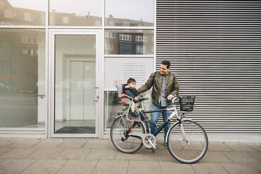 Lächelnder Vater auf dem Fahrrad mit seinem Sohn im Sicherheitssitz auf dem Bürgersteig in der Stadt - MASF17463