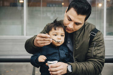 Vater füttert seinen Sohn mit Babynahrung, während er auf einer Bank in der Stadt sitzt - MASF17468