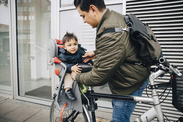 Father holding son sitting in safety seat of bicycle on sidewalk in city - MASF17472