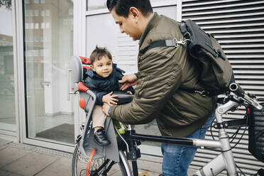 Vater hält seinen Sohn im Sicherheitssitz eines Fahrrads auf dem Bürgersteig in der Stadt - MASF17472