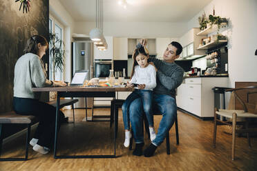 Father combing daughter's hair while mother working over laptop at home - MASF17477