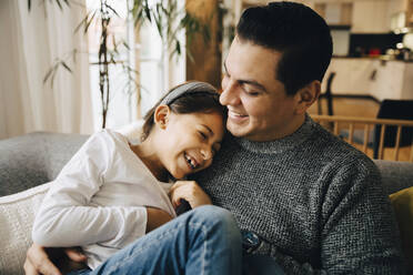 Smiling father and daughter spending leisure time while sitting on sofa at home - MASF17484