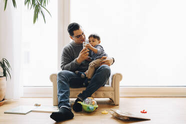 Full length of father playing with toddler son while sitting on sofa at home - MASF17485