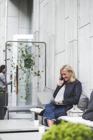 Lächelnde Geschäftsfrau, die mit dem Handy telefoniert und einen Laptop im Hof eines Kreativbüros benutzt, lizenzfreies Stockfoto