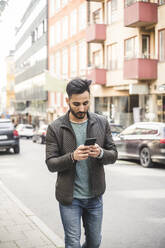 Junger Geschäftsmann, der ein Smartphone benutzt, während er auf dem Gehweg in der Stadt spazieren geht - MASF17492