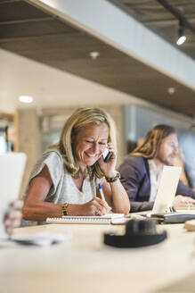 Lächelnde Geschäftsfrau, die in ein Tagebuch schreibt, während sie mit einem Smartphone im Büro telefoniert - MASF17509