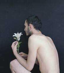 Side View Of Shirtless Young Man Holding Flower While Crouching Against Wall - EYF02662