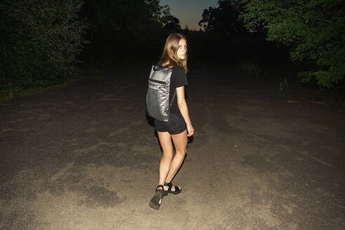 Rückansicht Porträt der jungen Frau zu Fuß auf dem Gehweg in der Abenddämmerung - EYF03024