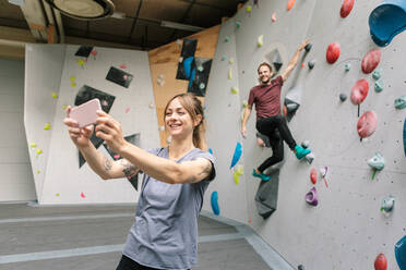 Lächelnde Frau nimmt Selfie durch Smartphone mit männlichen Freund Klettern Wand in der Turnhalle - MASF17517