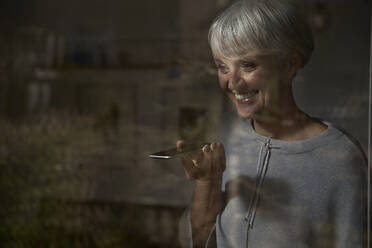 Porträt einer lächelnden älteren Frau hinter einer Fensterscheibe mit Smartphone - AUF00289