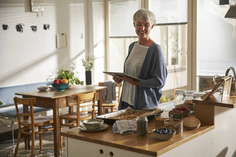 Porträt einer lächelnden älteren Frau, die mit einem digitalen Tablet in der Küche steht und Müsli zubereitet, lizenzfreies Stockfoto