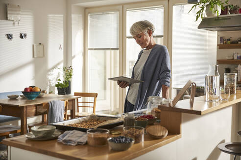 Senior woman standing in kitchen with digital tablet preparing granola - AUF00305