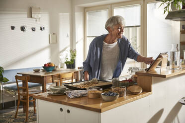 Lächelnde ältere Frau, die ein digitales Tablet in der Küche benutzt, während sie Müsli zubereitet - AUF00307