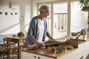 Smiling senior woman preparing granola looking at digital tablet - AUF00309