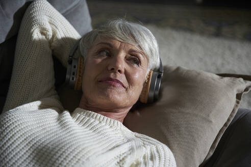 Porträt einer älteren Frau, die sich auf der Couch entspannt und mit Kopfhörern Musik hört - AUF00318
