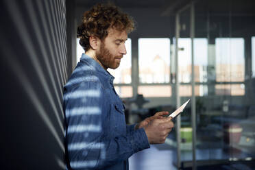 Casual businessman leaning against a wall in office using a tablet - RBF07249