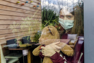 Portrait of girl with surgical mask and teddy bear behind window pane - SARF04513