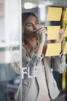 Businesswoman writing on adhesive notes on glass pane in office - RBF07344