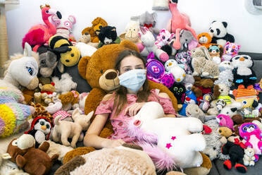 Girl with face mask, sitting on couch, covered in cuddly toys - SARF04516