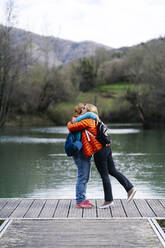 Two happy women with backpacks standing on jetty hugging each other, Valdemurio Reservoir, Asturias, Spain - DGOF00663